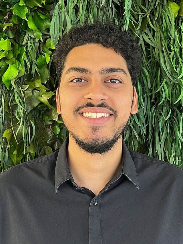 Smiling young man with curly black hair in front of a lush green wall.